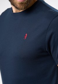 Tume sinine puuvillane T-särk ümarate varrukatega ja lühikeste varrukatega, mille vasakus rinnas on väike punane tikandiga logo.