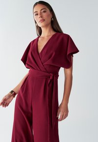 Mono marrón oscuro con mangas cortas y anchas, escote en V profundo, cintura cruzada con detalle de nudo y pantalones de pierna ancha a medida. Textura suave.