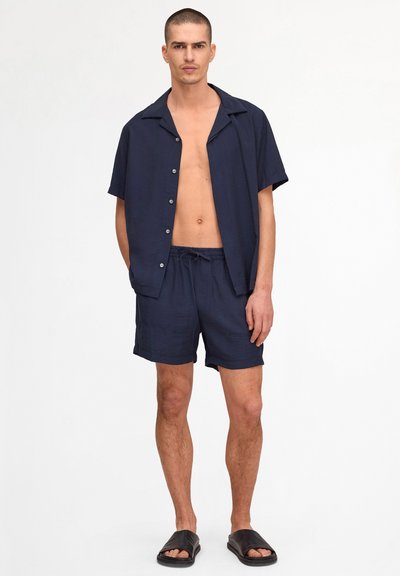 Man with short hair wearing unbuttoned navy short-sleeve shirt, matching navy shorts, and black slide sandals, standing against white background.