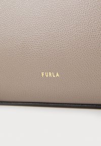 Teksturoidu vaaleanbeige nahkalaukku, jossa on kohokuvioitu "FURLA" -logo kullassa. Omistaa sileän pinnan ja hienovaraisen kuvion.