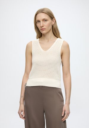 Femeie cu păr lung blond, purtând un top alb din tricot fără mâneci și pantaloni maro pe un fundal alb simplu.