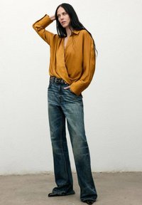 Blusa envolvente de manga larga en mostaza con puños anchos, combinada con jeans de mezclilla acampanados oscuros y un cinturón negro con tachuelas.