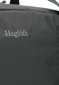 Zaino in tessuto nero con una trama a griglia, logo prominente "Haglöfs" ricamato in grigio, design semplice e spallacci regolabili.