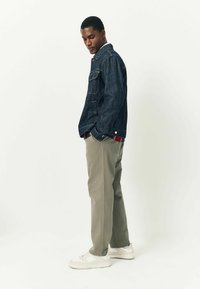 Denim jacket in dark blue with front pockets, featuring a metal button closure and contrasting red and white accents at the cuffs. Tan trousers and white sneakers.