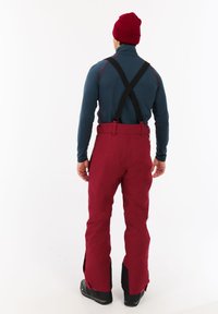 Rode sneeuwbroeken met zwarte schouderbanden, zijritsen en versterkte manchetten. Draagt over een blauwe longsleeve-top en een rode beanie.
