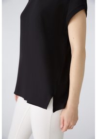Top nero a maniche corte con una vestibilità ampia, scollatura rotonda e spacchi laterali. Il tessuto sembra liscio e leggero, abbinato a pantaloni bianchi.
