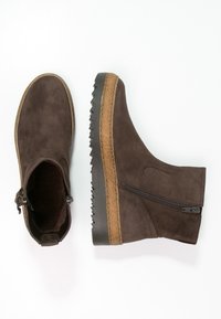 Brązowe zamszowe botki za kostkę z bocznym zamkiem, teksturowaną gumową podeszwą oraz kontrastującym jasnobrązowym akcentem u dołu. Okrągły krój noska.