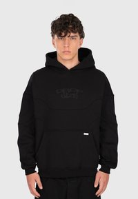 Junger Mann mit lockigem Haar, der einen schwarzen Hoodie mit Fronttasche und schwach eingesticktem Text "DROP SIZE" auf der Brust trägt, steht vor einem schlichten Hintergrund.