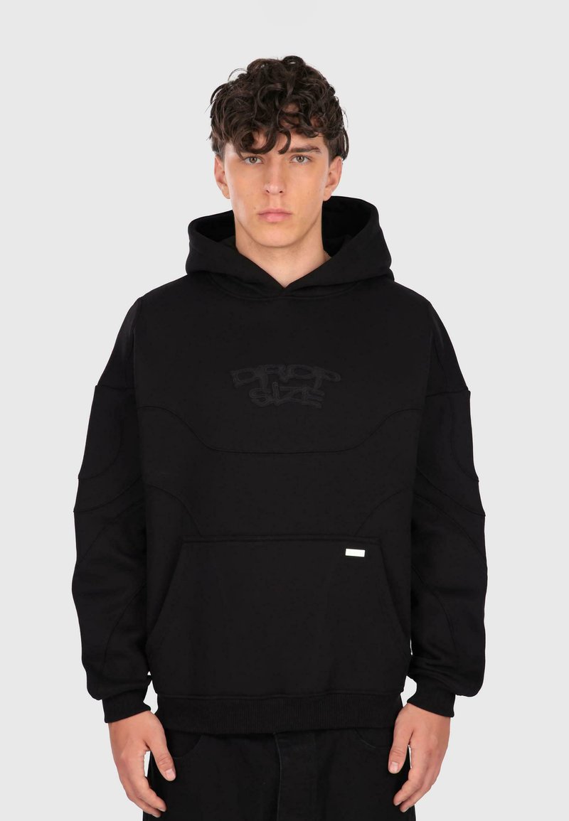 Junger Mann mit lockigem Haar, der einen schwarzen Hoodie mit Fronttasche und schwach eingesticktem Text "DROP SIZE" auf der Brust trägt, steht vor einem schlichten Hintergrund.