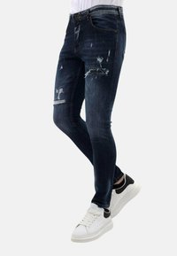 Donkerblauwe, versleten spijkerbroek met scheuren en een slim fit, gecombineerd met witte en zwarte sneakers. Gladde textuur, taps toelopend ontwerp.