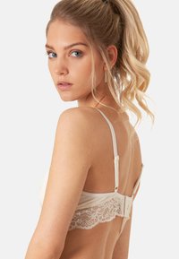 Sfiammante color beige chiaro con pizzo sul retro, spalline regolabili e chiusura a gancio. Tessuto liscio con un leggero luccichio.