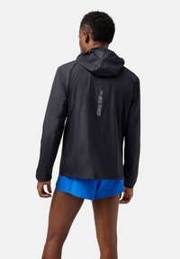 Giacca da corsa leggera nera con cappuccio, caratterizzata da un logo riflettente sulla schiena e tessuto liscio e resistente all'acqua. Abbinata a pantaloni corti blu.