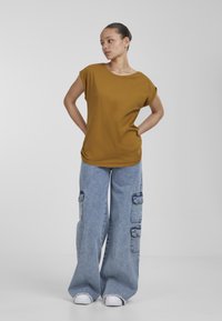 Senapsgul kortärmad t-shirt i kombination med vida blå denim cargobyxor med sidofickor och vita sneakers. Enkla bakgrund.