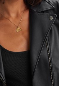 Złoty naszyjnik z okrągłym wisiorkiem o misternym wzorze, zawieszonym na cienkim łańcuszku, noszony na czarnej prążkowanej bluzce i skórzanej kurtce.