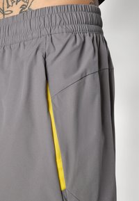 Grå atletiske shorts lavet af letvægtsstof, med en elastisk talje og en kontrasterende gul lynlåsdetailles på sidelommen.