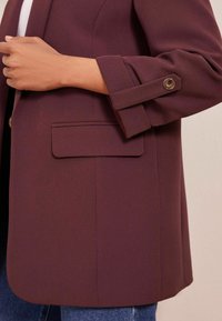 Bordový blazer s úzkým střihem, obsahující jednu kapsu, ohrnuté manžety upevněné knoflíkem a texturovanou tkaninu.