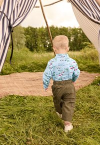 Camicia azzurro chiaro con un motivo giocoso di veicoli, abbinata a pantaloni cargo verde oliva e scarpe bianche. Erba e una tenda sullo sfondo.
