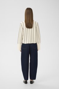 Femeie cu păr drept, maro, purtând un pulover crem cu dungi verticale negre și blugi evazați, bleumarin, stând cu spatele la privitor.