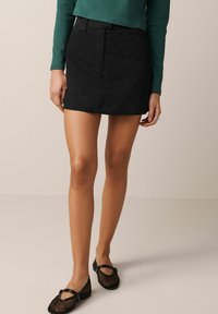 Femme portant une mini jupe noire, un haut à manches longues vert, et des ballerines Mary Jane en mesh noir, se tenant devant un fond beige uni.