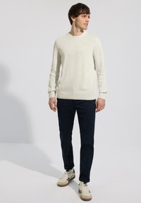 Homme portant un pull à manches longues beige clair, un pantalon bleu marine foncé et des baskets beiges, regardant vers la droite sur un fond blanc uni.