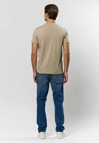 Mężczyzna stojący tyłem, ubrany w beżowy t-shirt, niebieskie dżinsy i białe trampki na jednolitym jasnym tle.