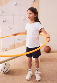 Ein Kind in einem weißen T-Shirt und navyblauen Shorts hält einen orangen Hula-Hoop-Reifen. Es trägt weiße Turnschuhe mit strukturierter Sohle und schwarze, gestreifte Socken.