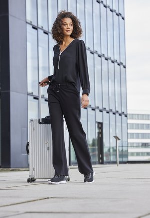 Schwarze Bluse mit langen Ärmeln und Reißverschlussdetails, schwarze Jogginghose mit Kordelzug, graue Sneaker und ein silberner Koffer auf einem gepflasterten Außenbereich in der Nähe von Glast Gebäuden.