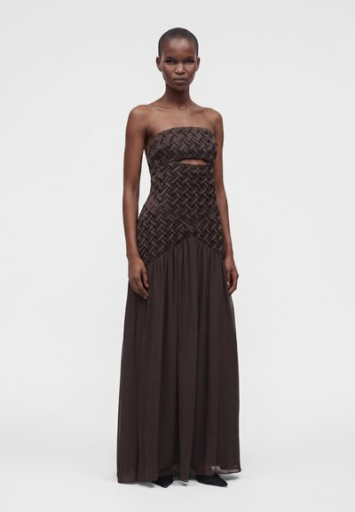 Γυναίκα που φορεί ένα strapless μακρύ σκούρο καφέ φόρεμα με πλεκτή λεπτομέρεια στο μπούστο και μικρό άνοιγμα κάτω από το στήθος, που στέκεται μπροστά σε απλό φόντο.