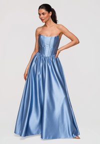 Femme portant une robe de bal en satin bleu clair sans bretelles, avec un corsage ajusté et une jupe ample, debout avec une main sur la hanche sur un fond uni.