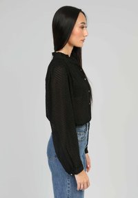Blouse noire transparente avec un motif texturé, col haut et fermeture à bouton. Manches longues ballon avec poignets élastiques ; associée à un jean taille haute.