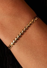 Bracelet en or arborant des maillons en forme de cœur avec une finition brillante, porté au poignet sur une peau texturée. Design simple et élégant.