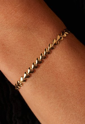 Gouden armband met hartvormige schakels en een glanzende afwerking, gedragen om een pols tegen een textuur van de huid. Eenvoudig en elegant ontwerp.