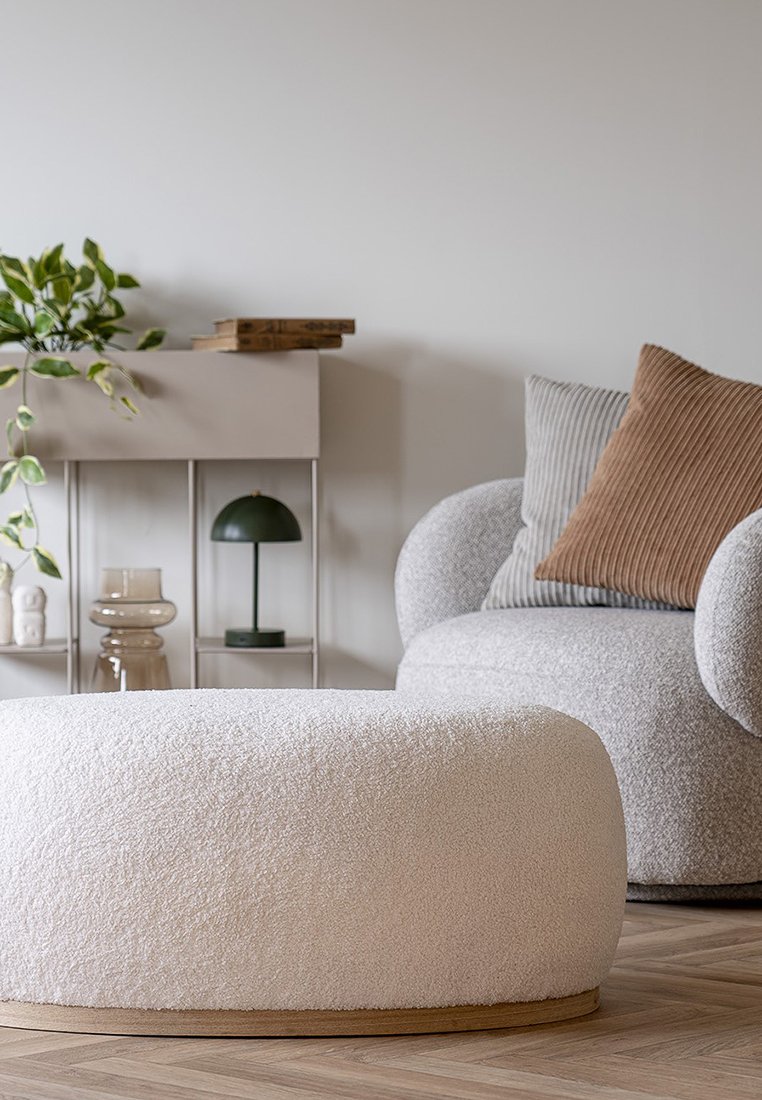 Runder Hocker aus weichem, cremefarbenem Textil mit Holzfuß. Umgeben von einem grauen Sofa, einer Pflanze und minimalistischer Dekoration auf einem hellen Holzboden.