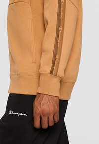 Felpa color sabbia con una texture liscia, caratterizzata da un nastro con logo lungo la manica e una tasca a marsupio. Abbinata a pantaloni neri.