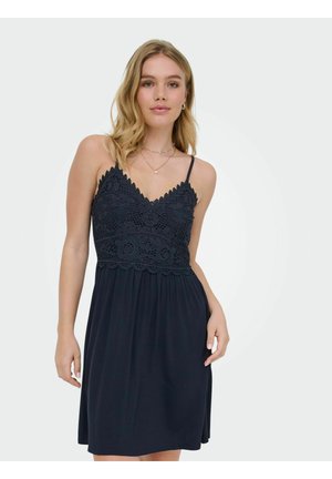 Kvinde iført en ærmeløs marineblå kjole med blonde detaljer på overdelen og tynde stropper, stående mod en enkel hvid baggrund.