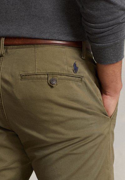 Primer plano de una persona que lleva pantalones verde oliva con un bolsillo trasero con botón y un cinturón de cuero marrón, con la mano metida en el bolsillo delantero.