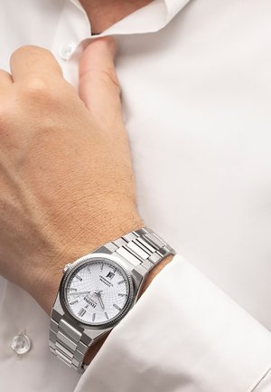 Left wrist wearing a silver metal watch with a white dial and date display, resting near the collar of a white dress shirt.