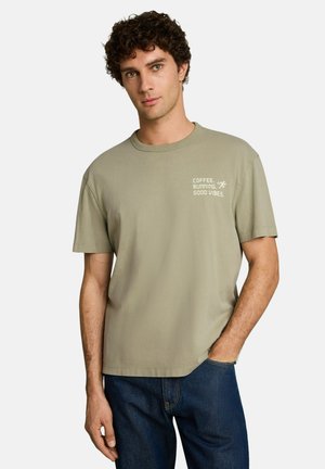 Mann mit lockigem Haar trägt ein hellolivgrünes T-Shirt mit dem Text "COFFEE, RUNNING, GOOD VIBES" und posiert mit einer Hand in der Tasche einer dunkelblauen Jeans.
