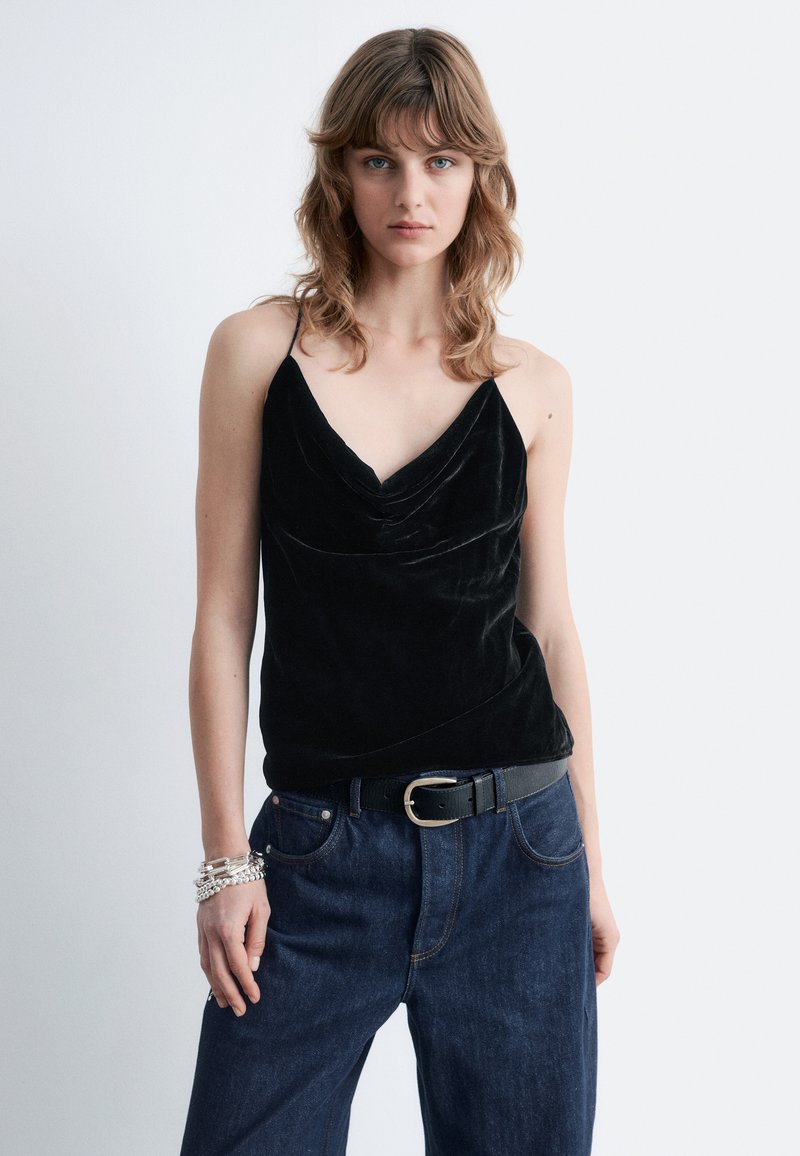 Black velvet camisole with a cowl neckline, thin straps, paired with high-waisted dark denim jeans and a black belt. Silver bracelets on wrist.