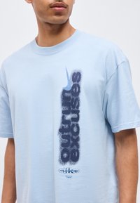 Άνδρας που φοράει ανοιχτό μπλε μπλουζάκι Nike με κατακόρυφο σκούρο μπλε κείμενο "SB Dunk" και λογότυπο Swoosh στο μπροστινό μέρος.