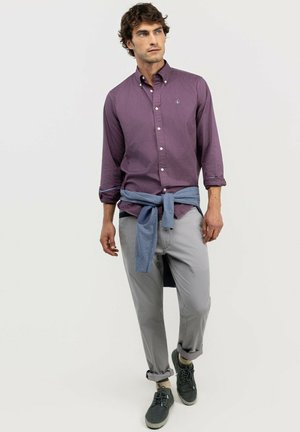 Hombre con camisa morada abotonada, pantalones gris claro, zapatillas grises y un suéter azul atado alrededor de la cintura, de pie frente a un fondo liso.