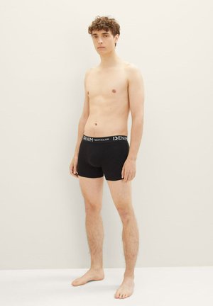 Schwarze Baumwoll-Boxershorts mit einem weißen Bund, auf dem "DENIM TOM TAILOR" steht. Das Modell steht barfuß vor einem hellen Hintergrund.