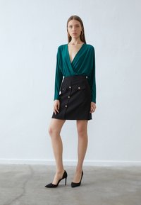 Haut enroulé vert à manches longues, associé à une mini-jupe noire avec des poches avant et des boutons dorés. Porté avec des talons aiguilles noirs.