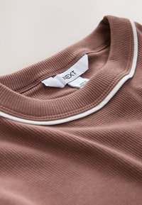 Tela marrón acanalada con borde blanco en el cuello, con una etiqueta que dice "NEXT." Textura suave con un ajuste relajado.