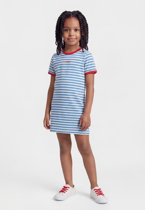 Dievča stojí, má na sebe modro-biele pruhované šaty s červeným lemovaním a biele tenisky s červenými šnúrkami, na jednoduchom pozadí.