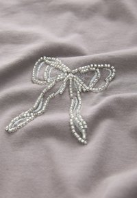 Tela gris con un diseño de lazo con cuentas en plata y cuentas transparentes, con una textura suave y un bordado detallado que delimita la forma.