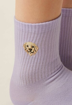 Fliederfarbige, gerippte Socke, die am Knöchel getragen wird, mit einem kleinen, bestickten Golden Retriever-Hundegesicht in der Nähe der Manschette.