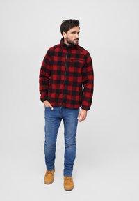 Rød- og sortternet fleecejakke med høj krave, lynlås og brystlomme, parret med blå jeans og brune støvler.