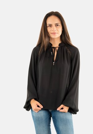 Donna con lunghi capelli castani che indossa una blusa nera a maniche lunghe con dettagli in pizzo e jeans blu, in piedi su uno sfondo bianco uniforme.