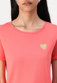 Koralinė trumpa rankovė, pagaminta iš minkštos medžiagos, su apvaliu kaklu ir auksiniu blizgučio širdelės logotipu kairėje krūtinės pusėje.